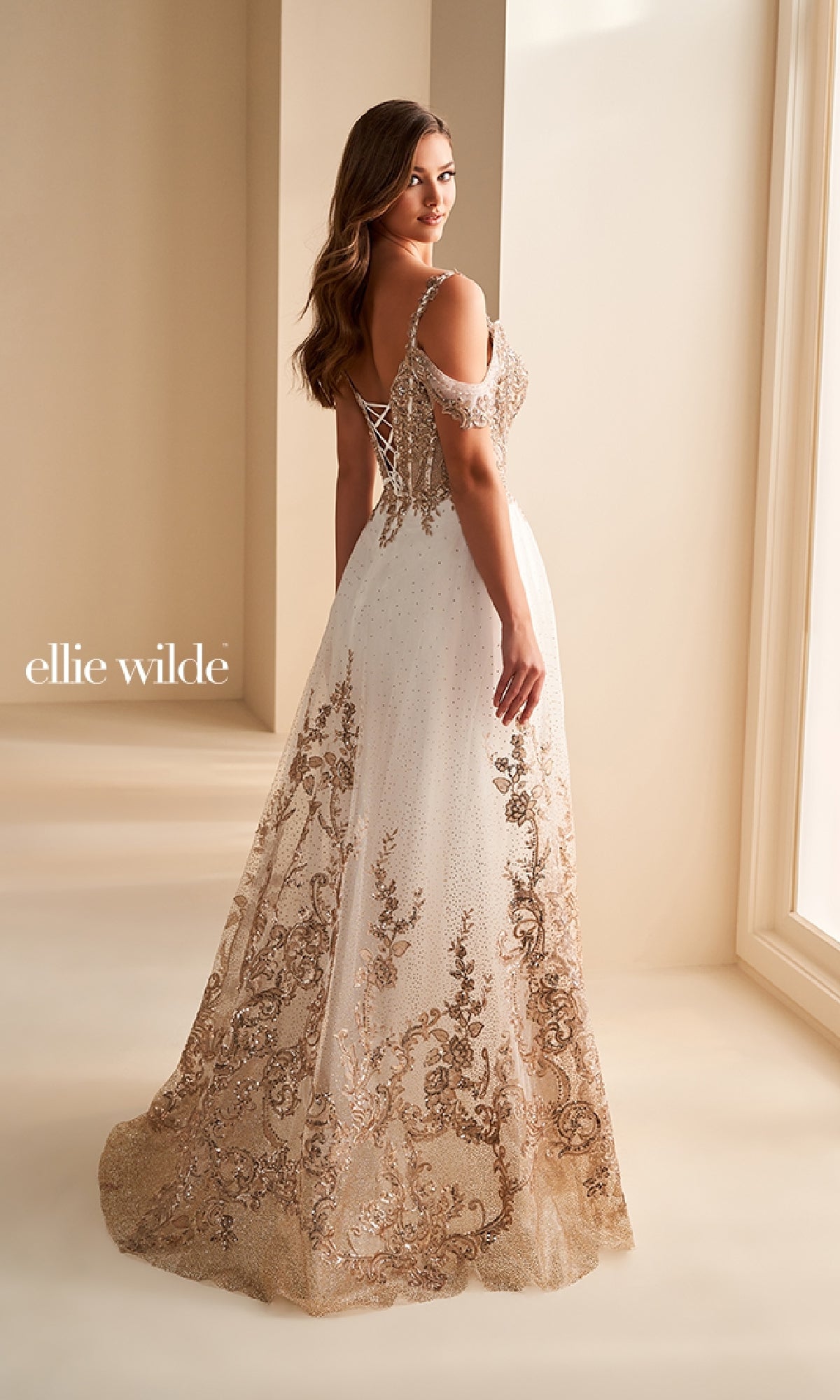 A woman stands near a window in the Ellie Wilde EW36220 long prom dress—a white and gold gown with lace-up back, glitter tulle, and floral accents. The image also features the text